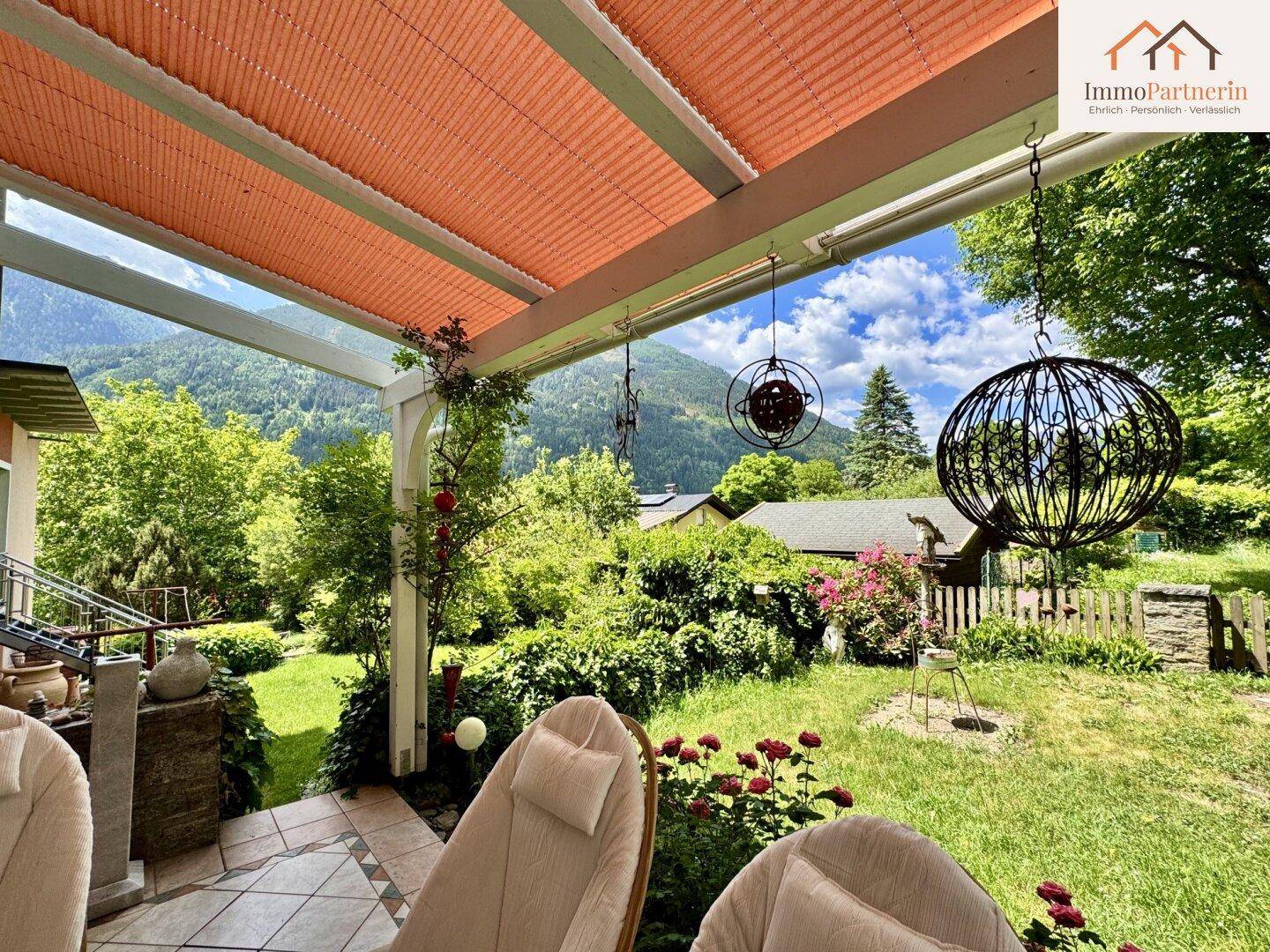 Überdachte Terrasse mit bequemen Liegestühlen und Blick auf den grünen Garten und die Berge.