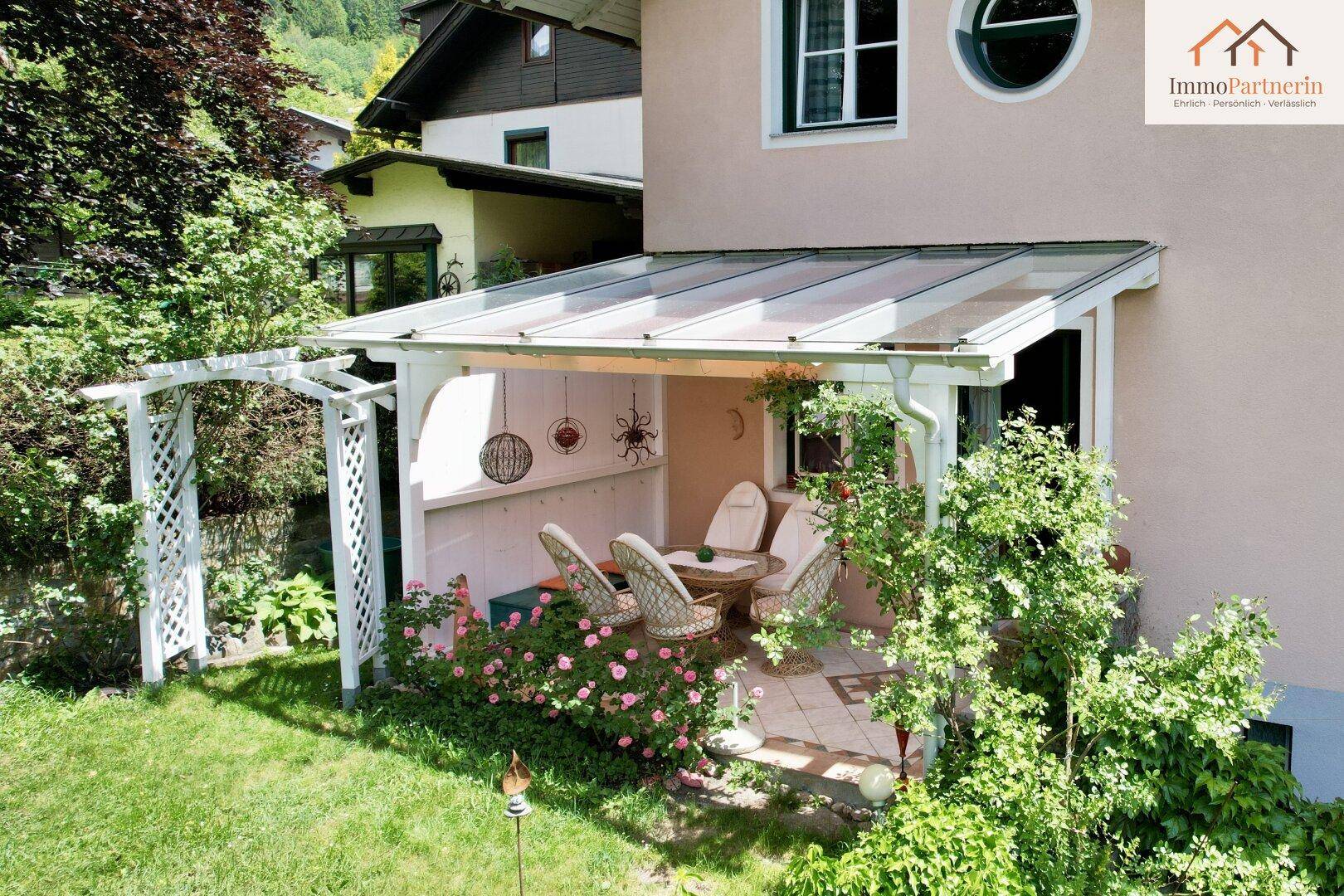 Überdachte Terrasse mit Sitzgelegenheiten, umgeben von blühenden Pflanzen und Grünflächen.