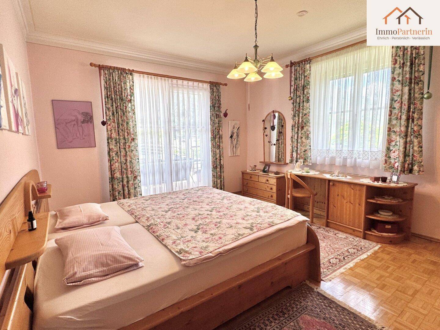 Schlafzimmer mit Doppelbett, Holzmöbeln, floralen Vorhängen und Zugang zum Balkon.