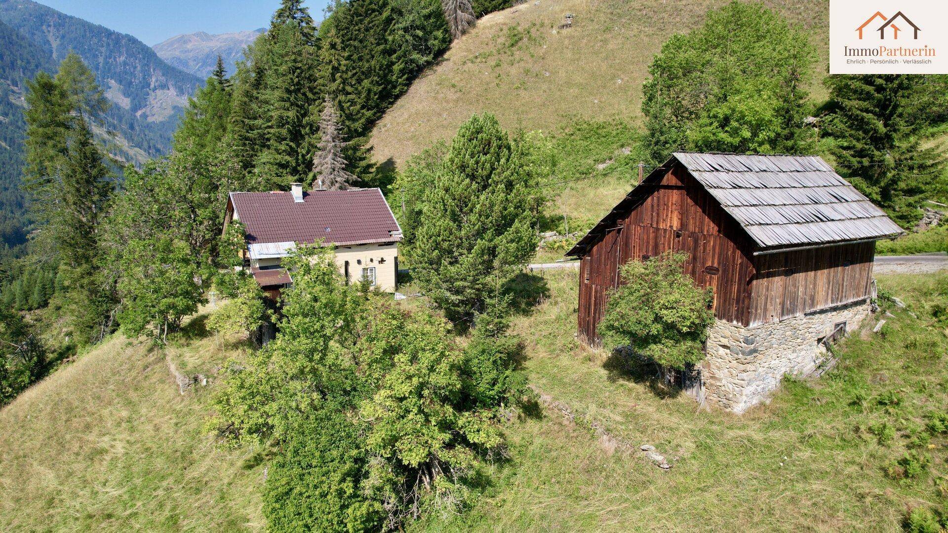 Zwei ländliche Gebäude in einer bergigen Landschaft mit viel Grün und Wald.