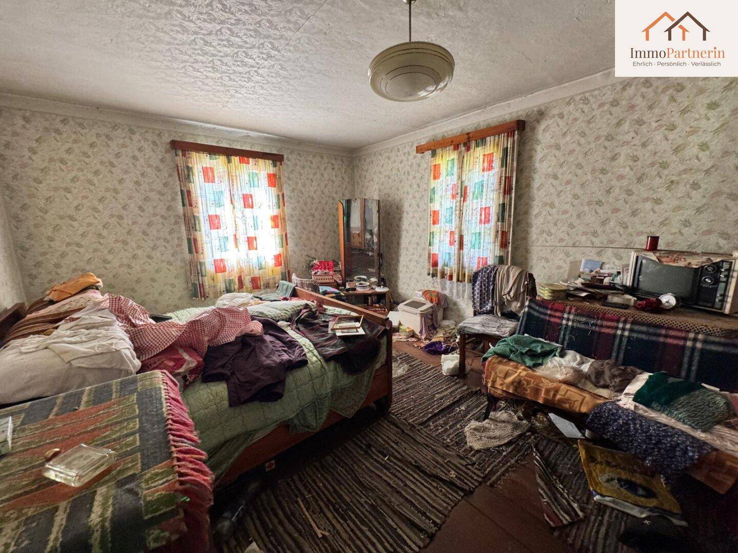 Unordentliches Schlafzimmer mit alten Möbeln, gemusterter Tapete und zwei Fenstern.