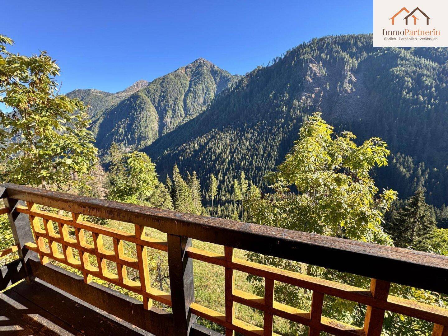 Balkon mit rustikalem Holzgeländer und atemberaubendem Blick auf die bewaldeten Berge unter blauem Himmel.