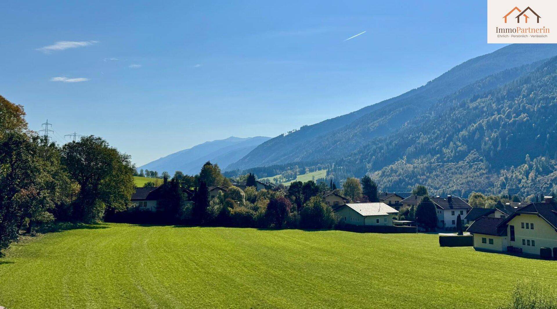 Panoramablick über eine grüne Wiese auf eine Siedlung und die majestätische Bergkette im Hintergrund.