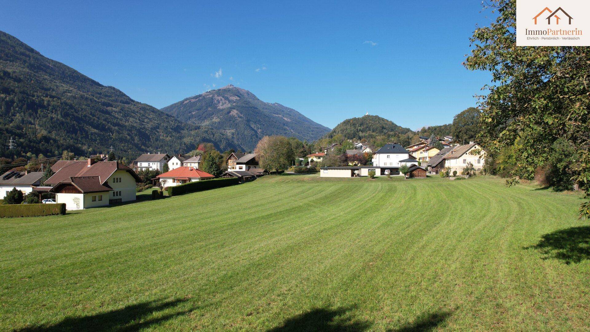 Idyllische Landschaft mit weitläufiger Wiese, umgeben von Wohnhäusern und imposanten Bergen.