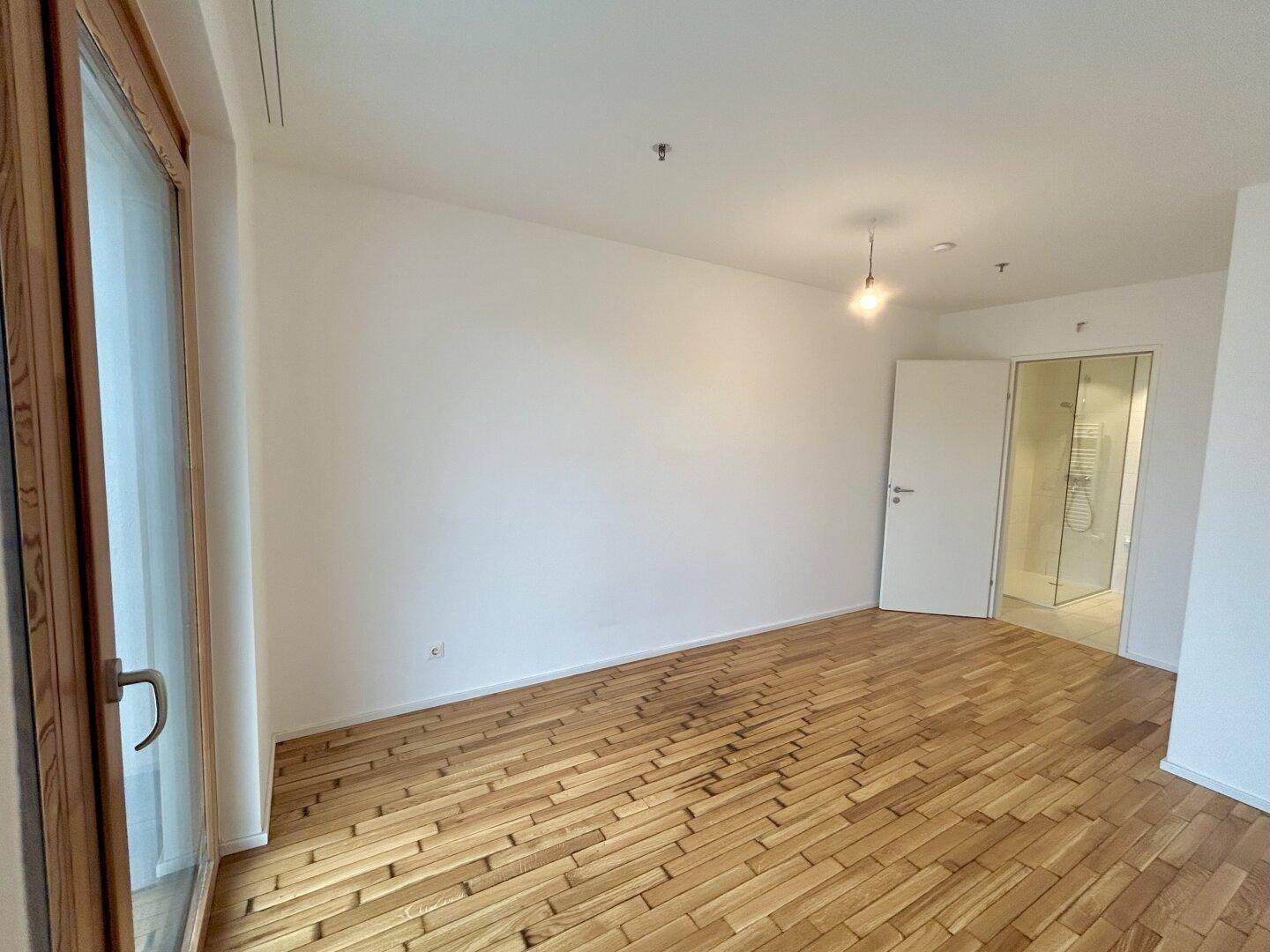 Helles Schlafzimmer mit Parkettboden, großem Fenster und direktem Zugang zum modernen Badezimmer.