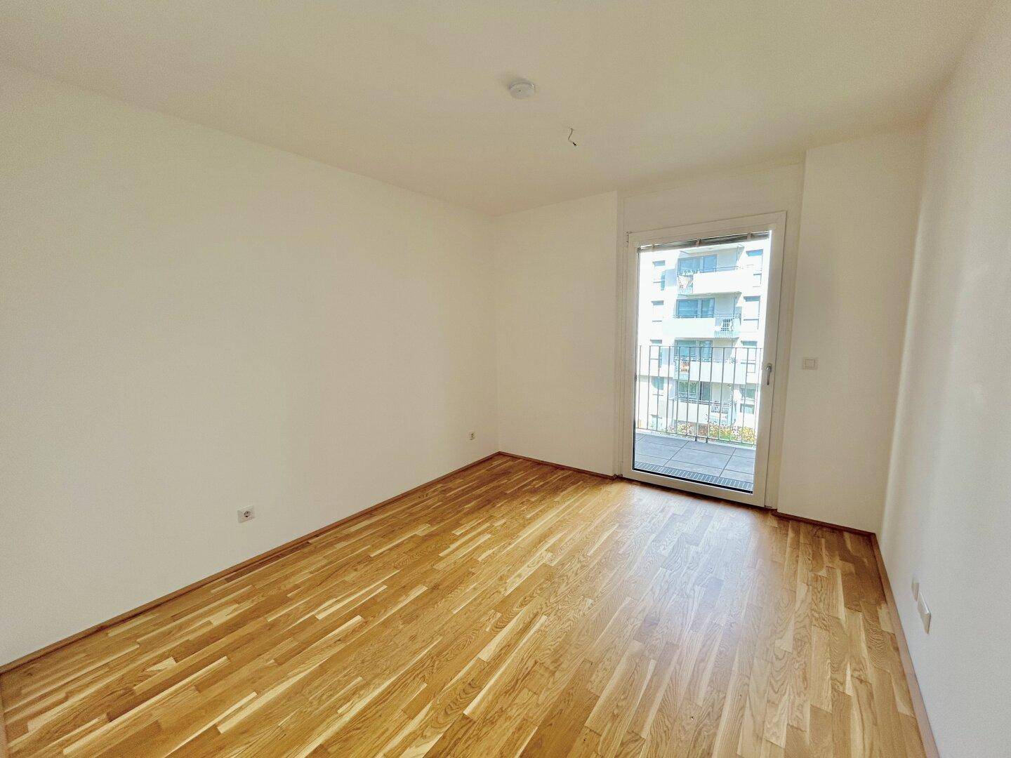 Helles Schlafzimmer mit Parkettboden und Zugang zu einem Balkon, in neuwertigem Zustand.