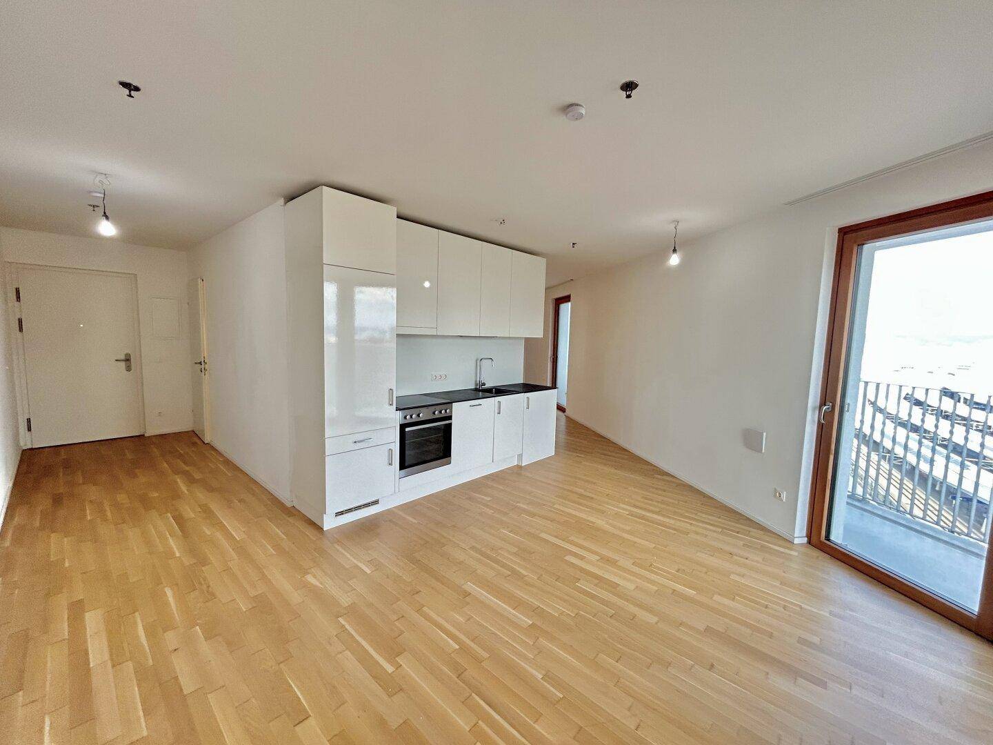 Offener Wohnbereich mit integrierter Küchenzeile, Parkettboden und Zugang zu einem Balkon.