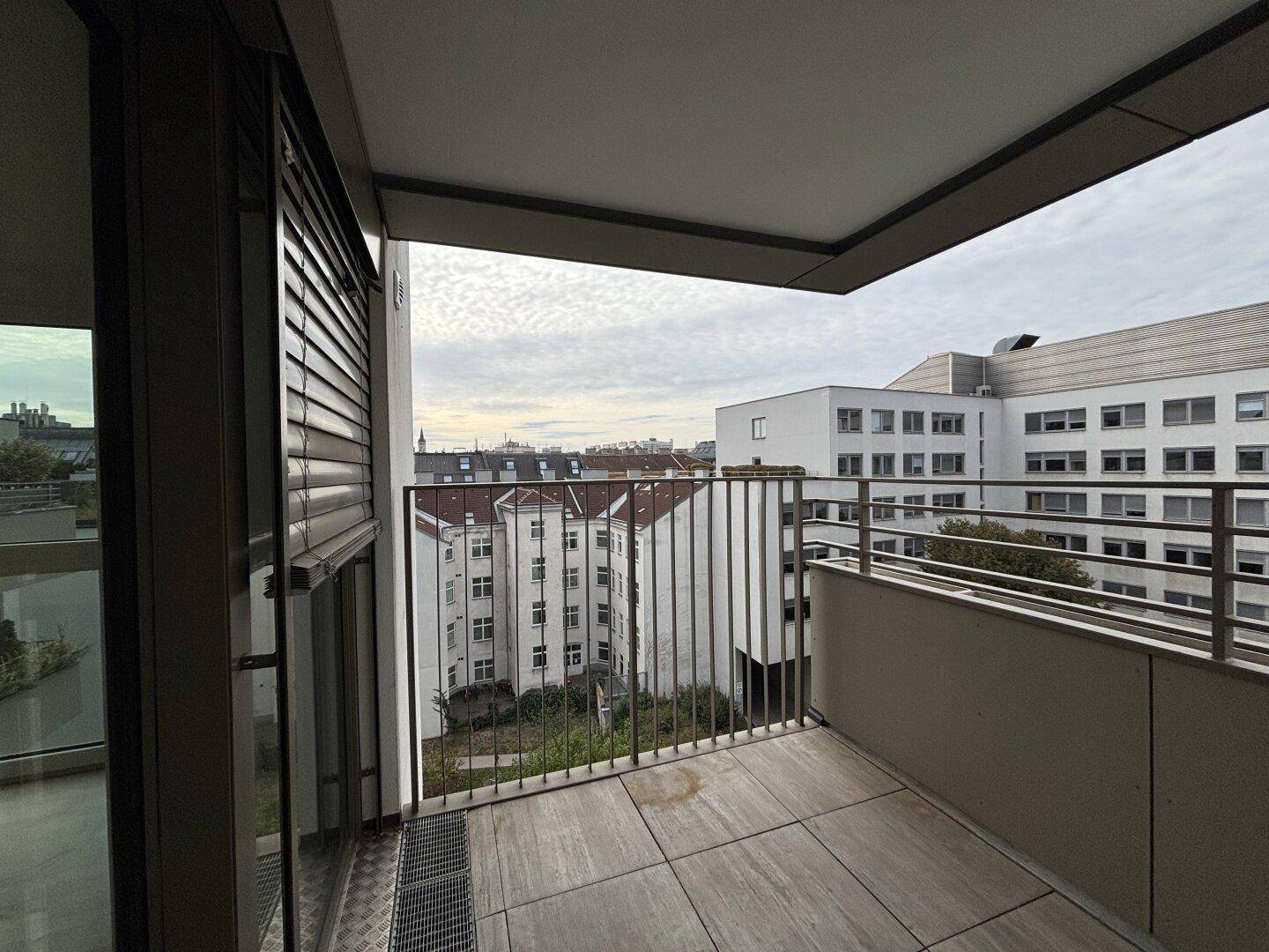 Balkon mit Fliesenboden und Blick auf die städtische Bebauung, mit Rollladen am Fenster.