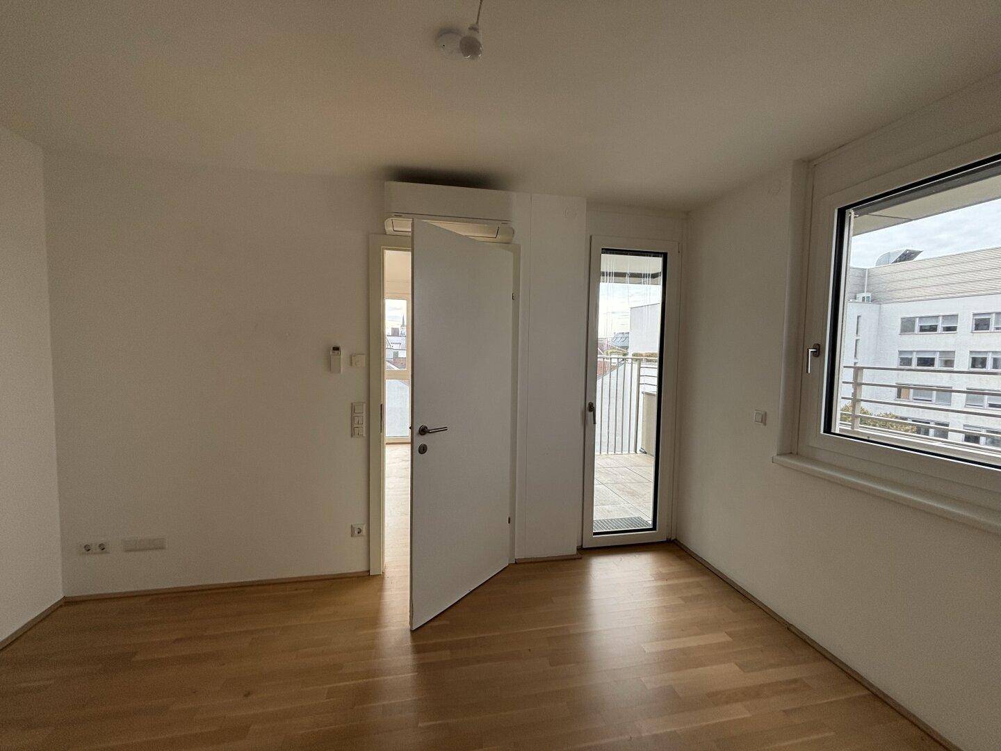 Helles Schlafzimmer mit Parkettboden, Klimaanlage und Zugang zu einem kleinen Balkon.