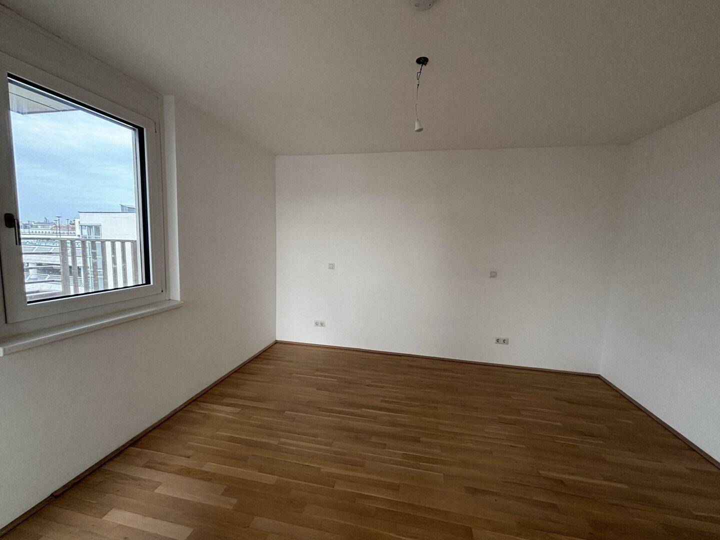 Leeres Zimmer mit hellem Parkettboden und großem Fenster, das viel Tageslicht hereinlässt.