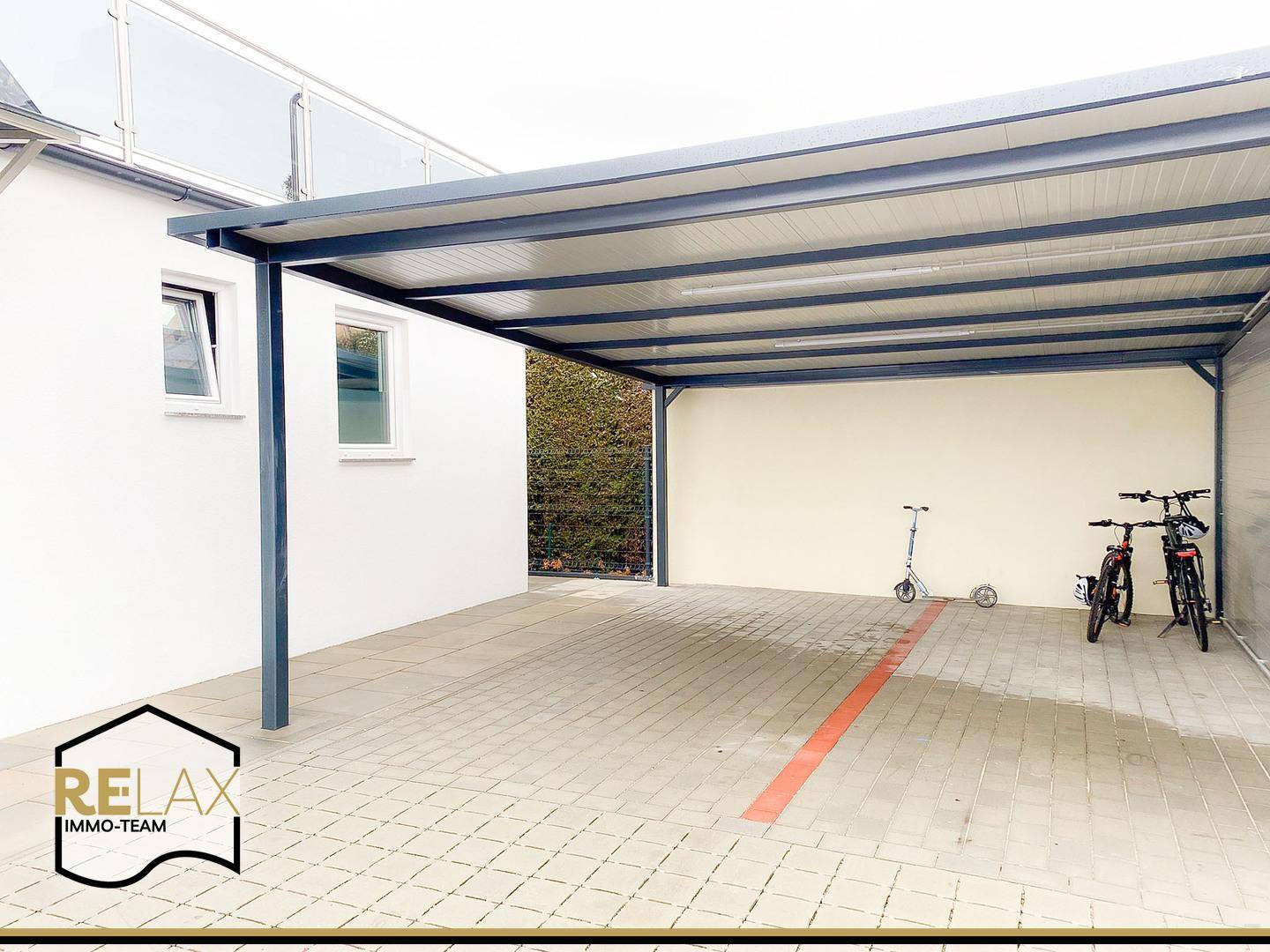Überdachter Carport mit gepflastertem Boden, bietet Platz für Fahrzeuge und Fahrräder.