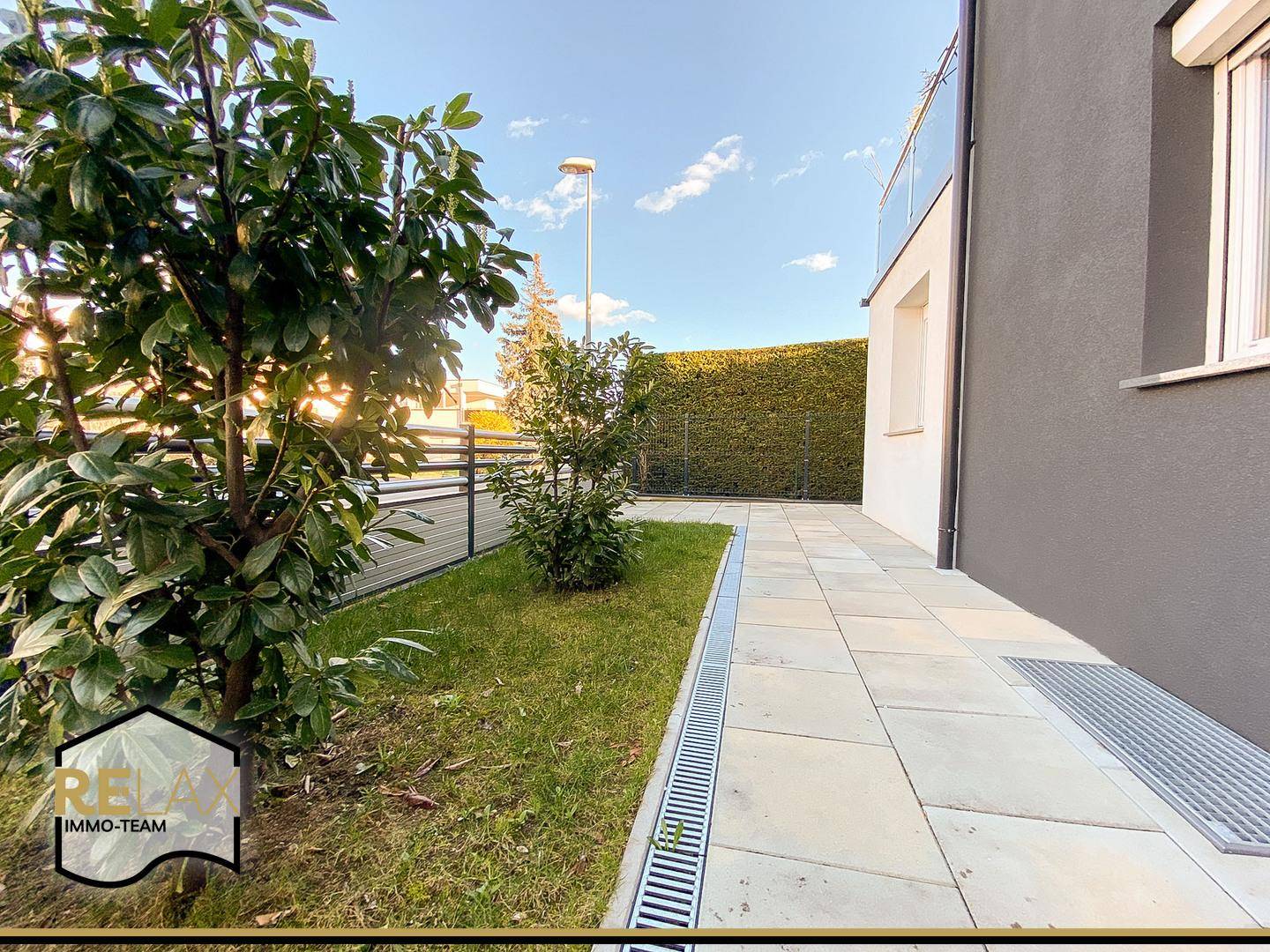 Kleiner Gartenbereich mit Rasenfläche und gepflastertem Weg neben dem Haus, umgeben von Hecken.