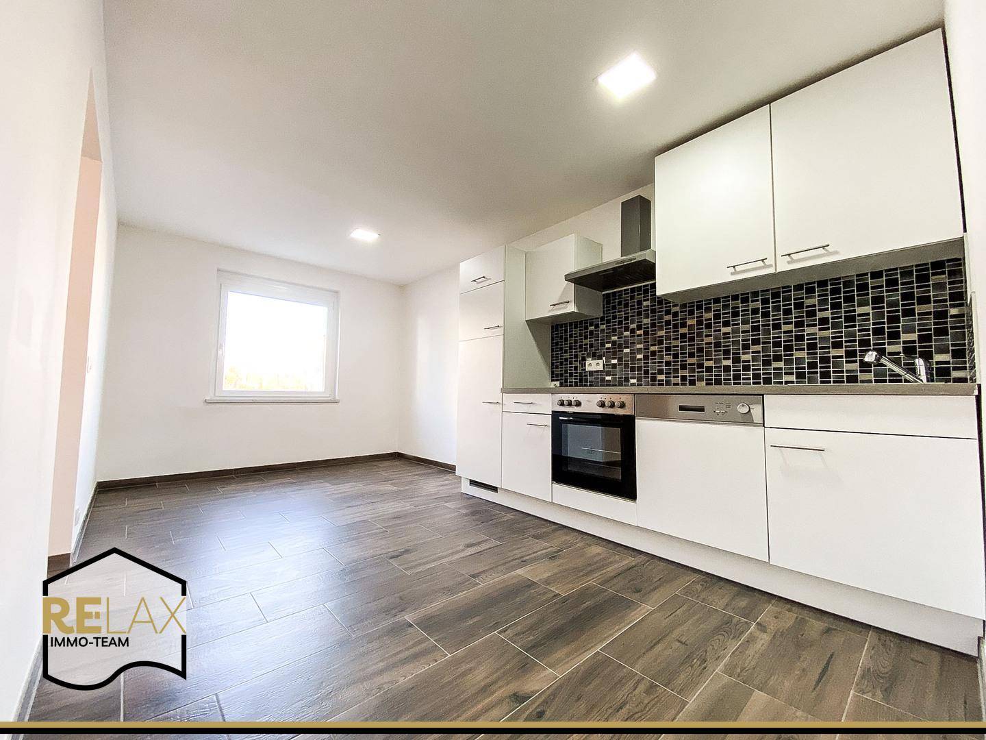 Moderne, unmöblierte Küche mit weißen Schränken, dunklem Fliesenspiegel und Holzoptik-Fliesenboden.