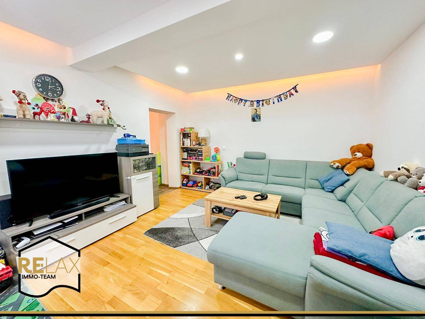 Gemütliches Wohnzimmer mit Ecksofa, Holzboden und moderner Deckenbeleuchtung, ideal für Familien.