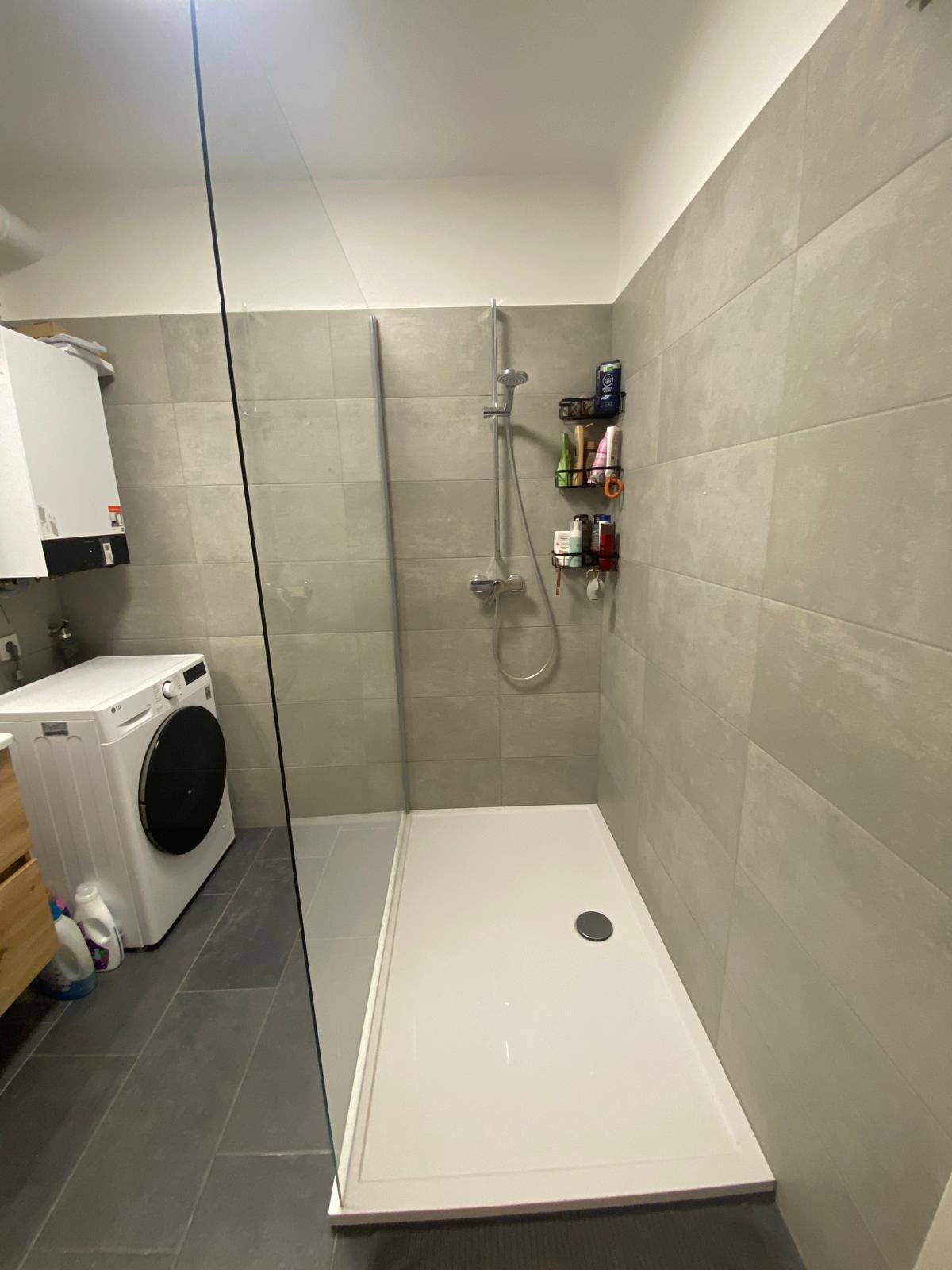 Modernes Badezimmer mit ebenerdiger Dusche, grauen Fliesen und einer Waschmaschine.