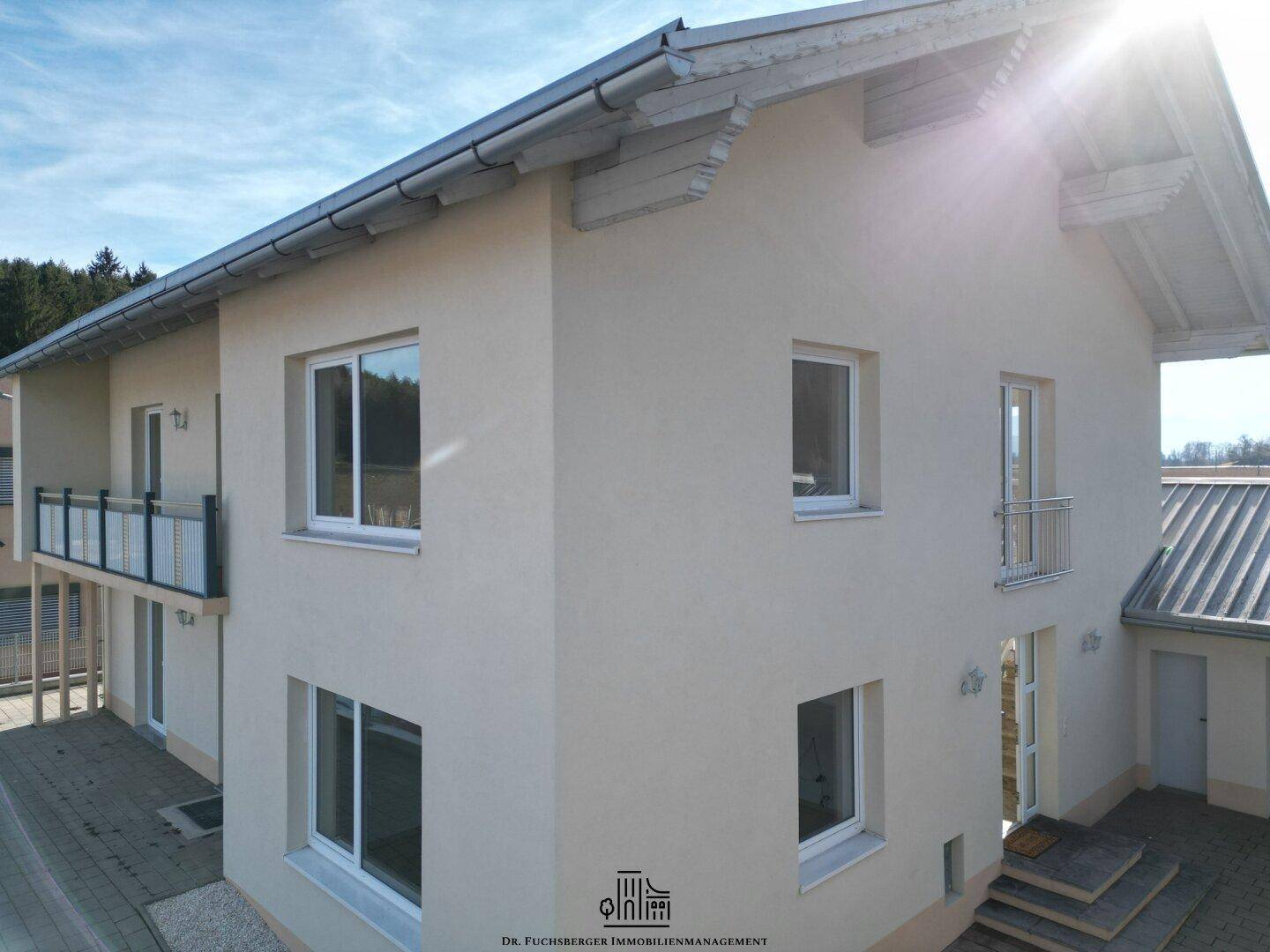 Modernes Wohnhaus mit heller Fassade, Balkonen und gepflastertem Vorplatz unter blauem Himmel.