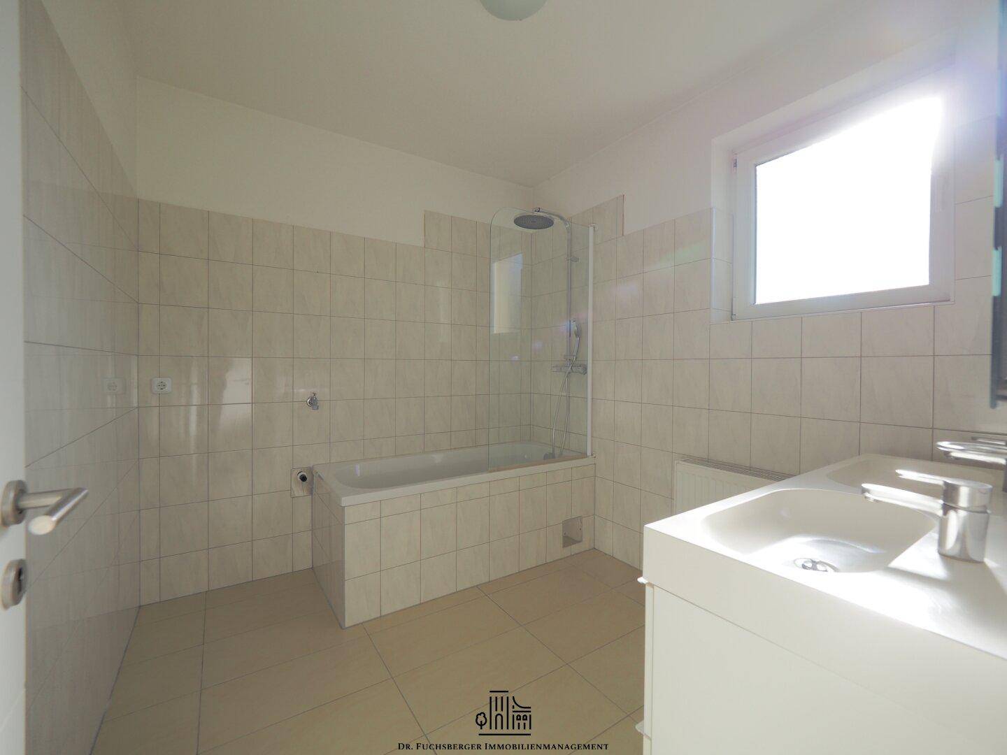 Modernes Badezimmer mit Badewanne, Dusche und Waschtisch, hell gefliest und mit Fenster.