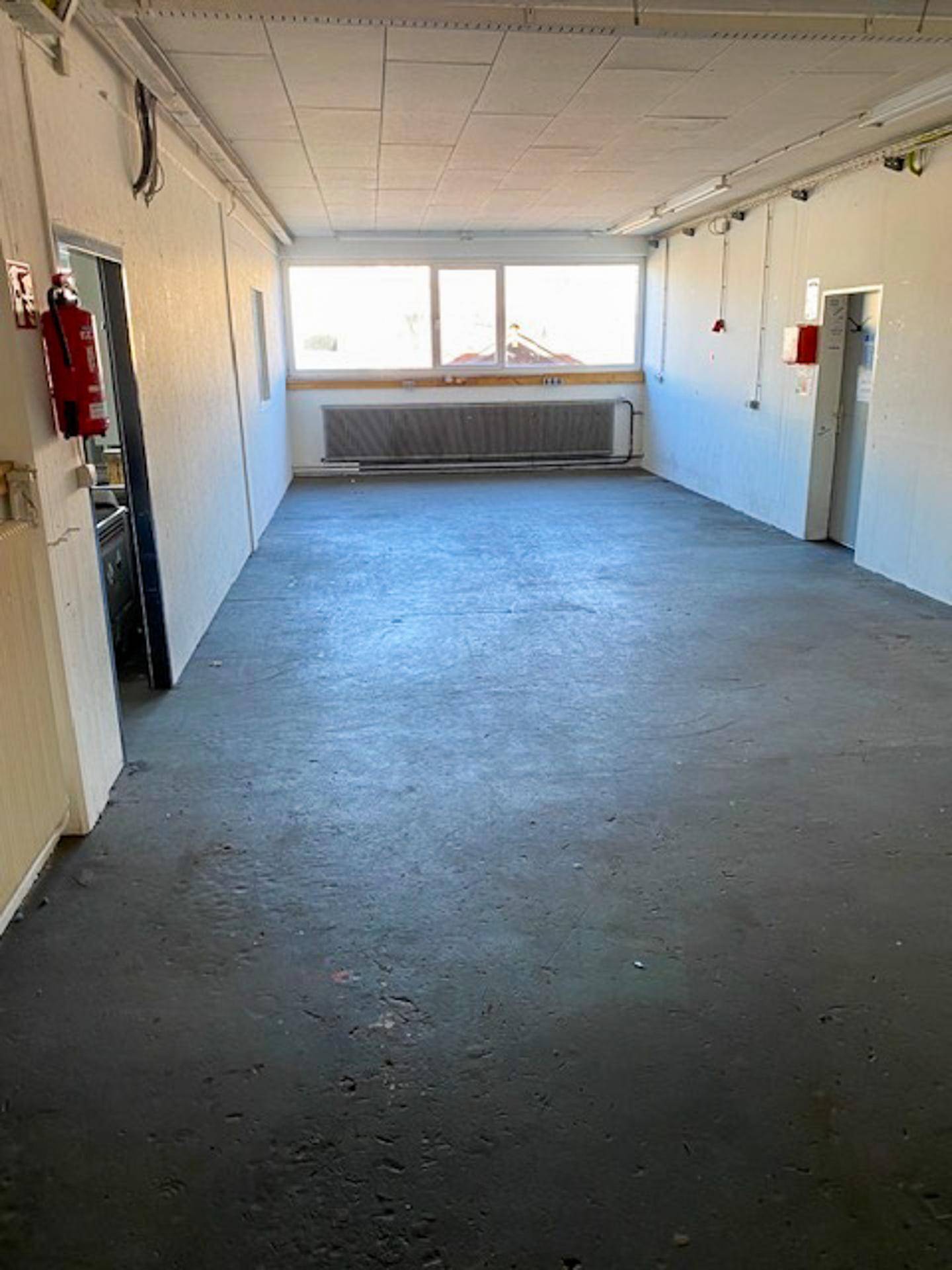 Leerer Raum mit Betonboden, einem großen Heizkörper unter dem Fenster und einfacher Beleuchtung.