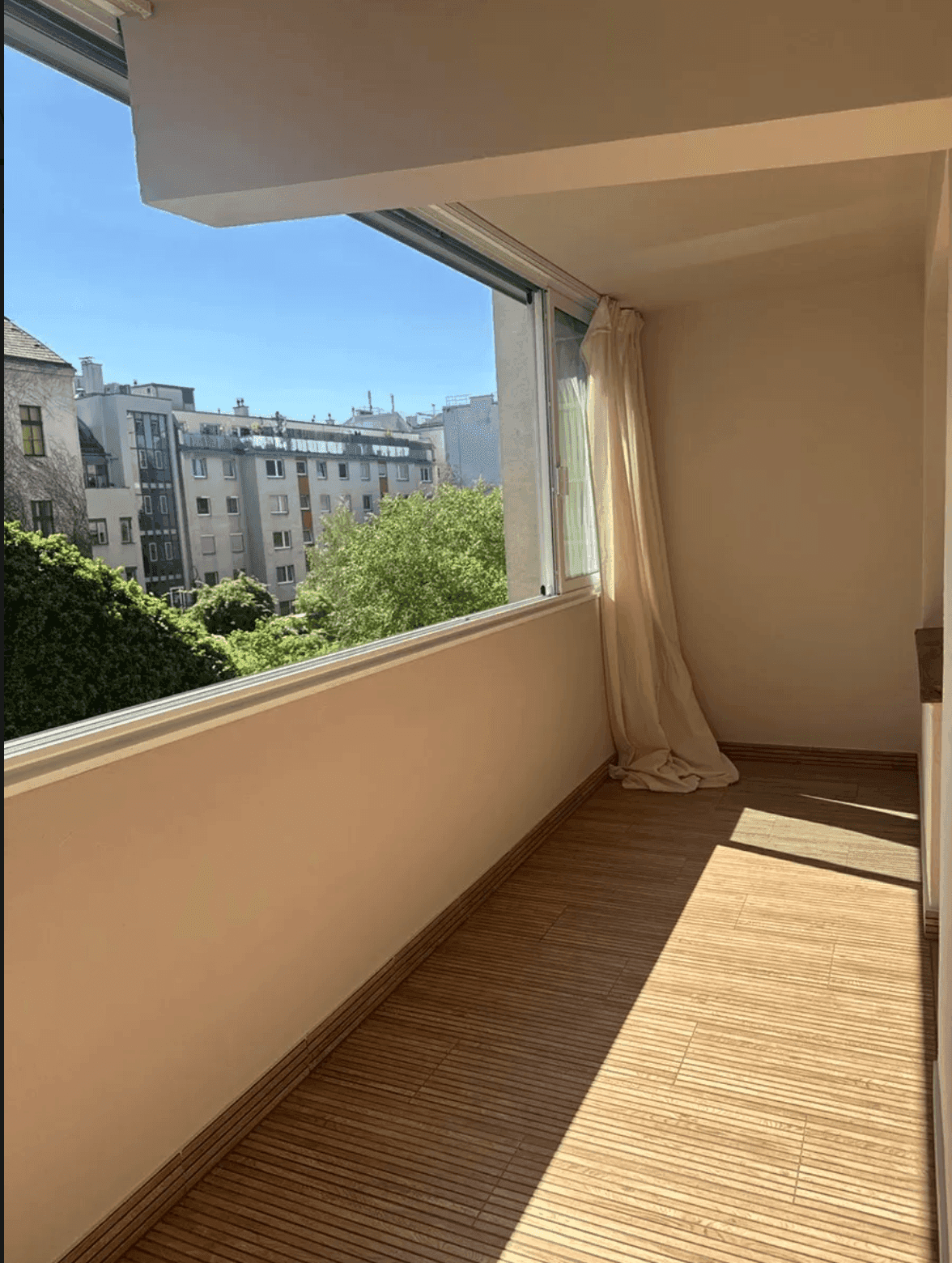 Verglaster Balkon mit Holzbodenfliesen und Blick auf die umliegenden Stadtgebäude und Bäume.