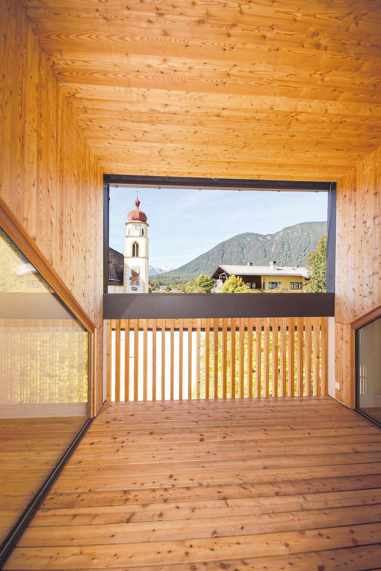 Überdachter Balkon mit Holzboden und weitem Blick auf die Berge und ein Dorf mit Kirchturm.