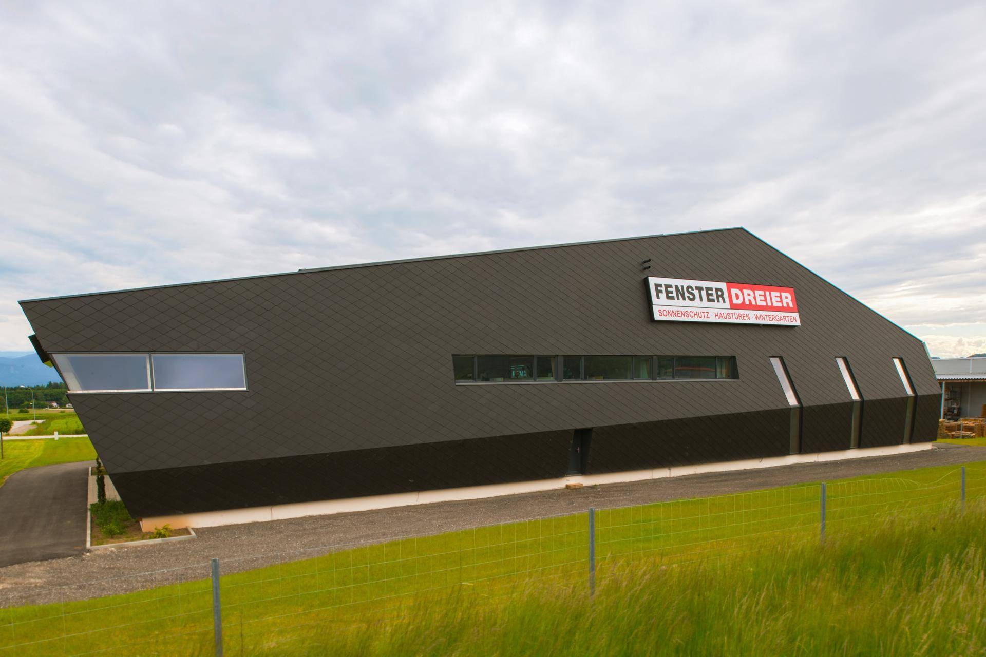 Moderne Geschäftsfassade mit dunklen Schindeln und horizontalen Fenstern, umgeben von grünen Feldern.
