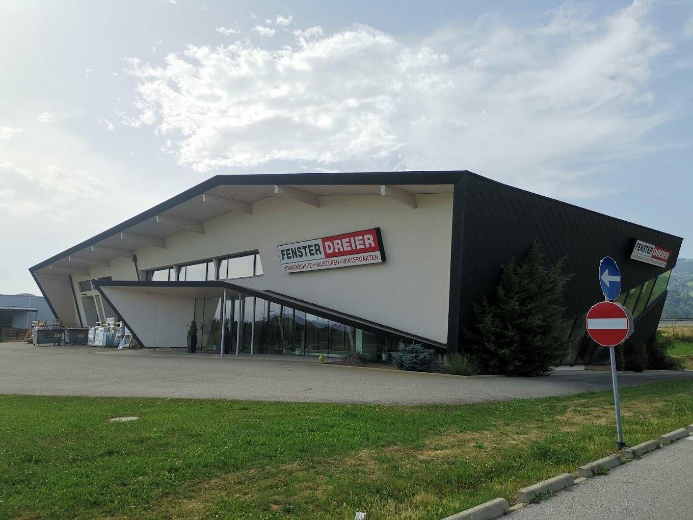 Modernes Geschäftsgebäude mit markanter Architektur, großen Glasflächen und Firmenlogo unter bewölktem Himmel.