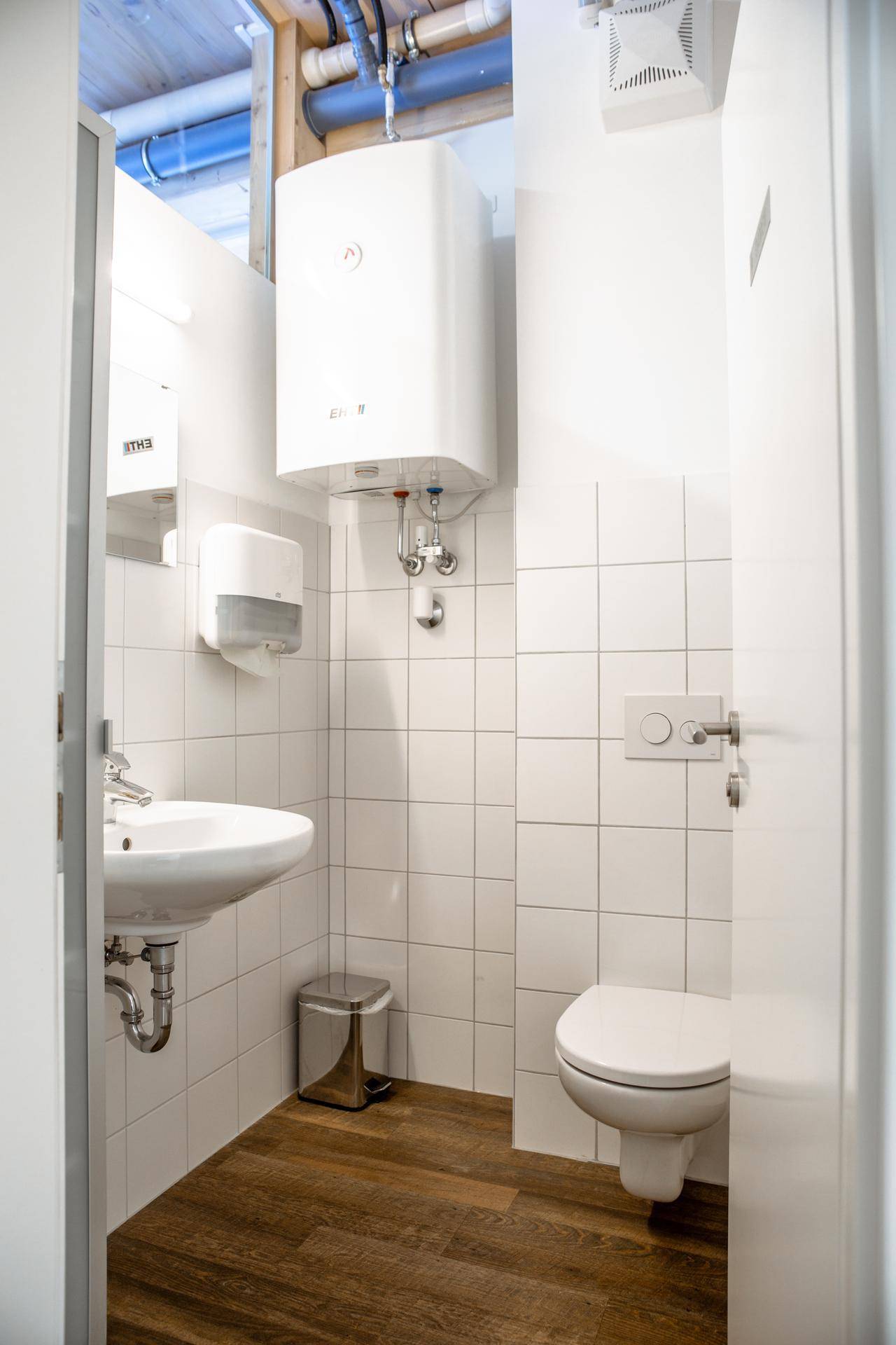 Saubere Toilette mit weißen Fliesen, Waschbecken und Warmwasserbereiter, mit Holzboden und sichtbaren Rohren.