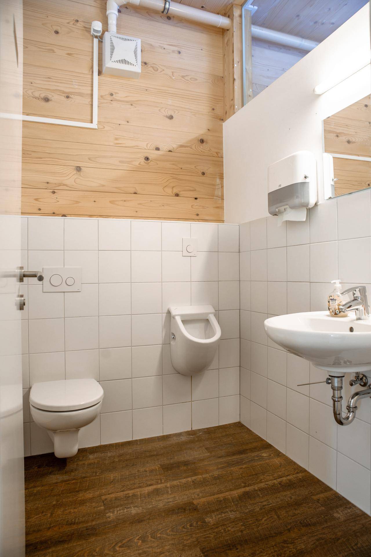 Moderne Toilette mit weißen Fliesen, Waschbecken und Urinal, kombiniert mit Holzwänden und -boden.