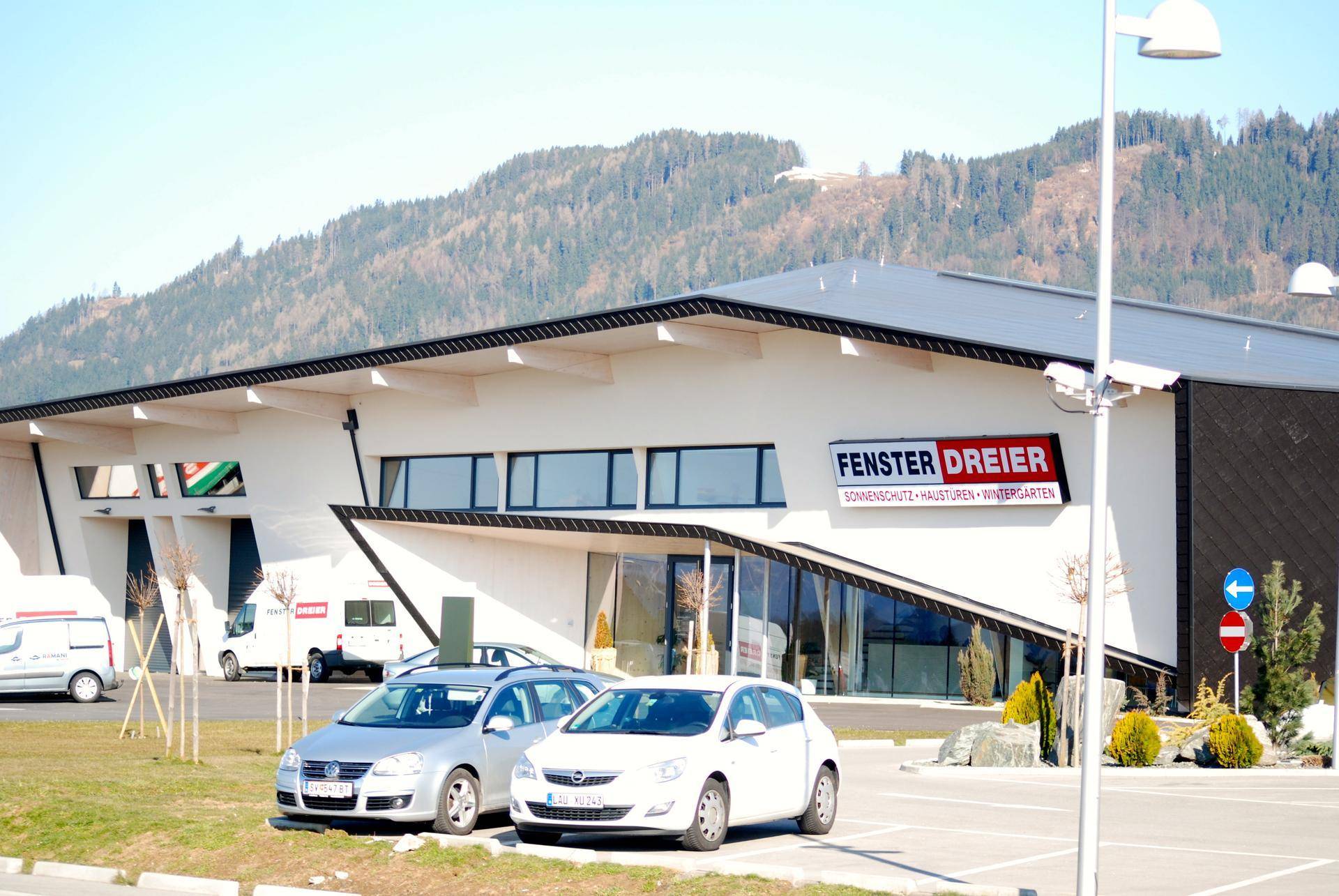 Modernes Geschäftsgebäude mit heller Fassade und großem Parkplatz, umgeben von grünen Hügeln.