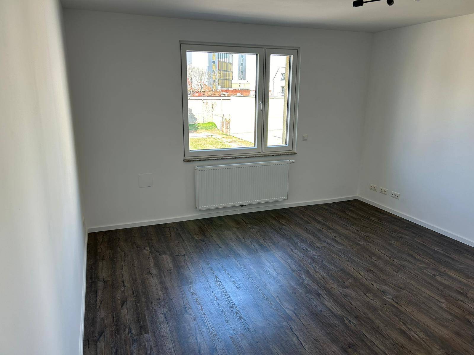 Helles Zimmer mit Laminatboden und Blick auf einen grünen Außenbereich.