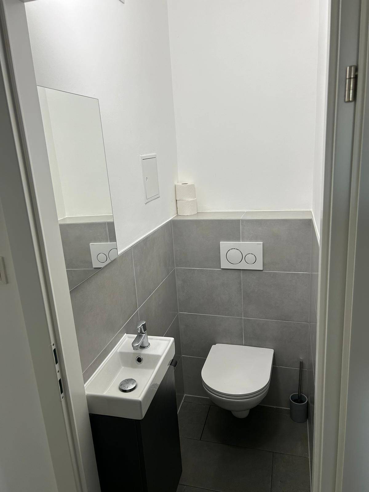 Modernes Gäste-WC mit grauen Fliesen, wandhängendem WC und kleinem Waschbecken.