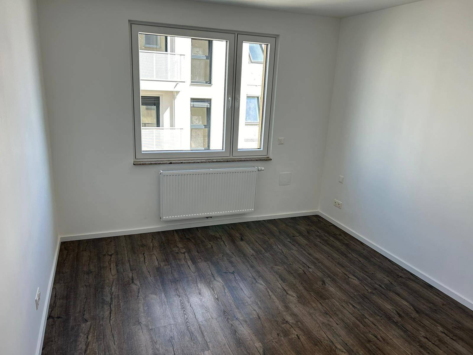 Leeres Zimmer mit dunklem Laminatboden und Fenstern mit Blick auf ein anderes Gebäude.