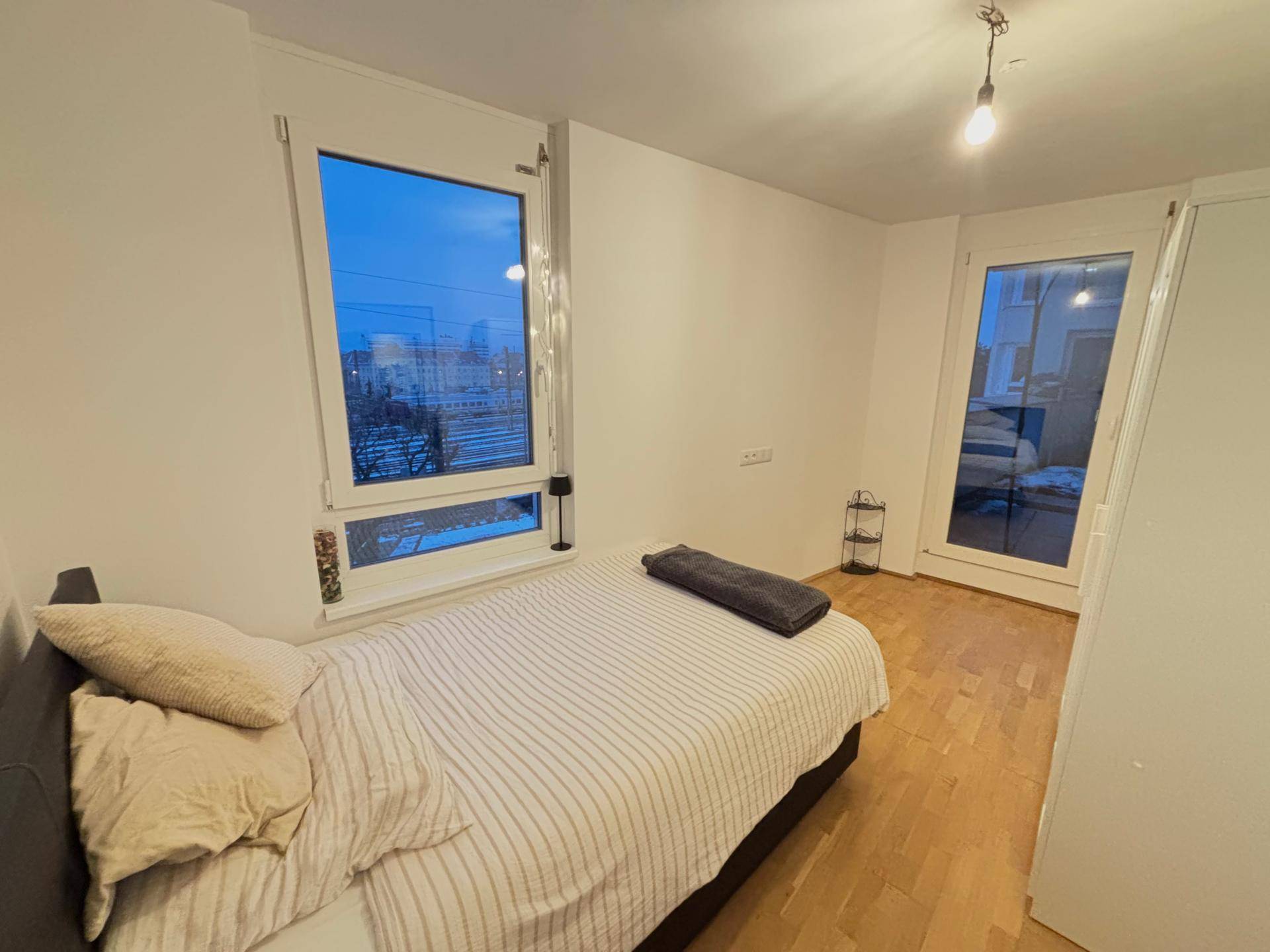 Schlafzimmer mit großen Fenstern, Holzboden und Zugang zu einem möglichen Balkon oder Außenbereich.