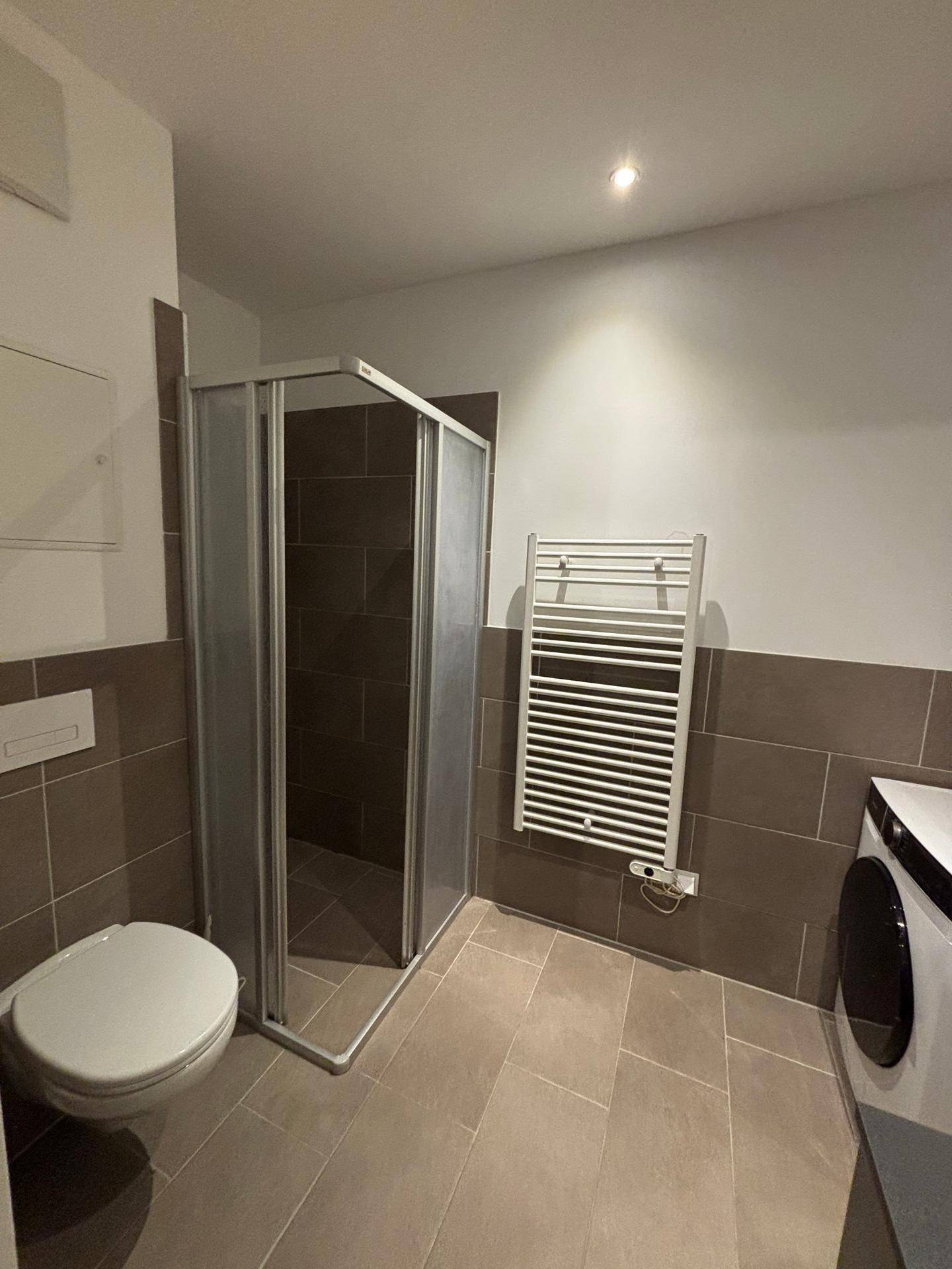 Modernes Badezimmer mit Duschkabine, Wand-WC, Fliesenboden und beheiztem Handtuchhalter.