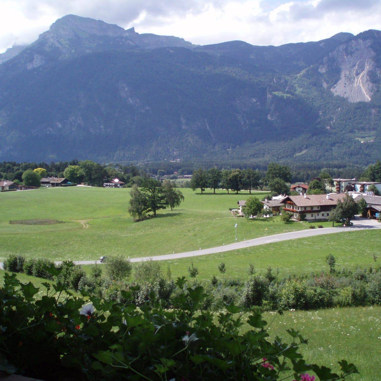 Weitläufiger Blick über grüne Wiesen und eine ländliche Siedlung mit majestätischen Bergen im Hintergrund.