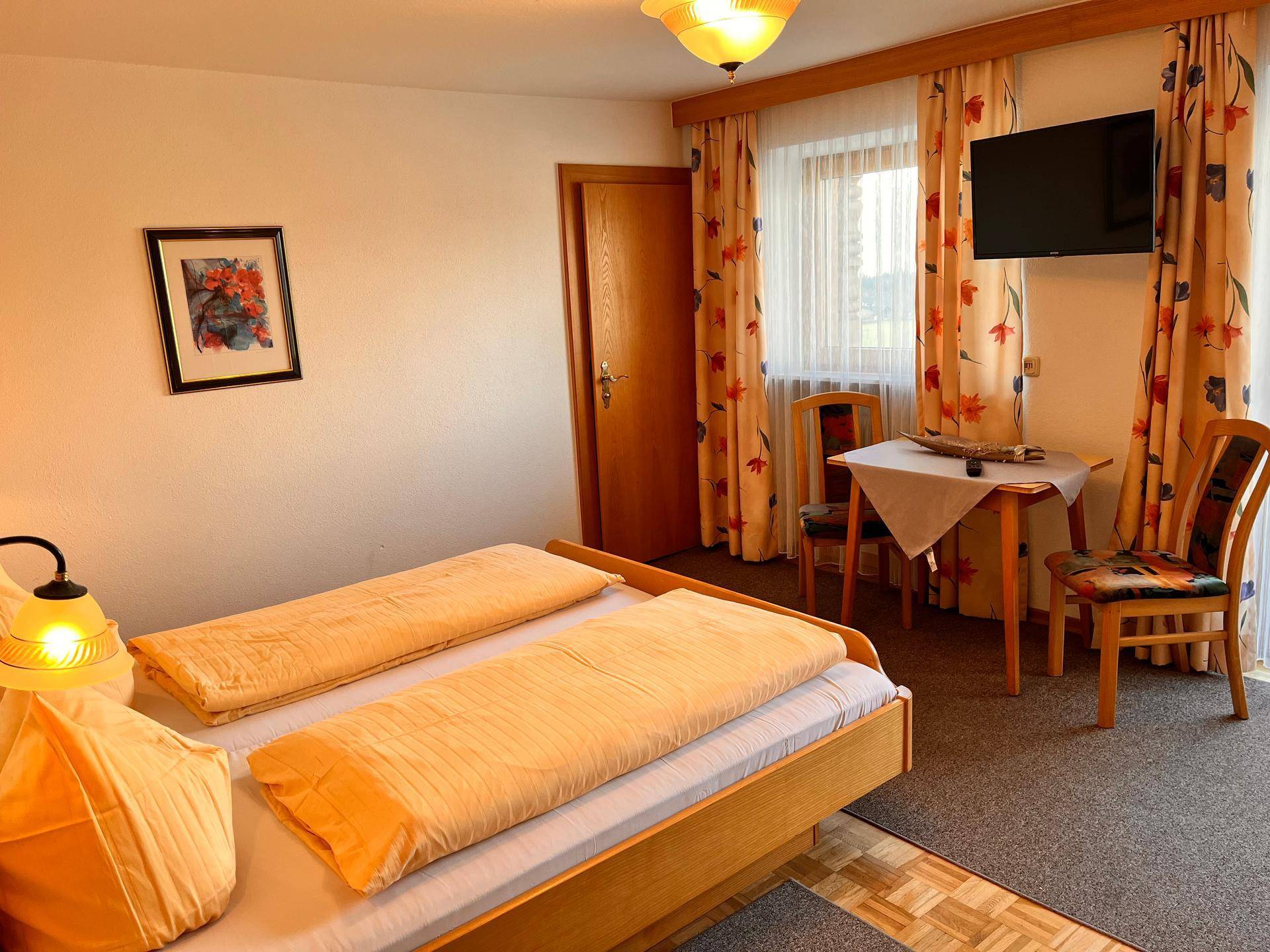 Helles Schlafzimmer mit floralen Vorhängen, Fernseher und kleinem Tisch mit Stühlen.