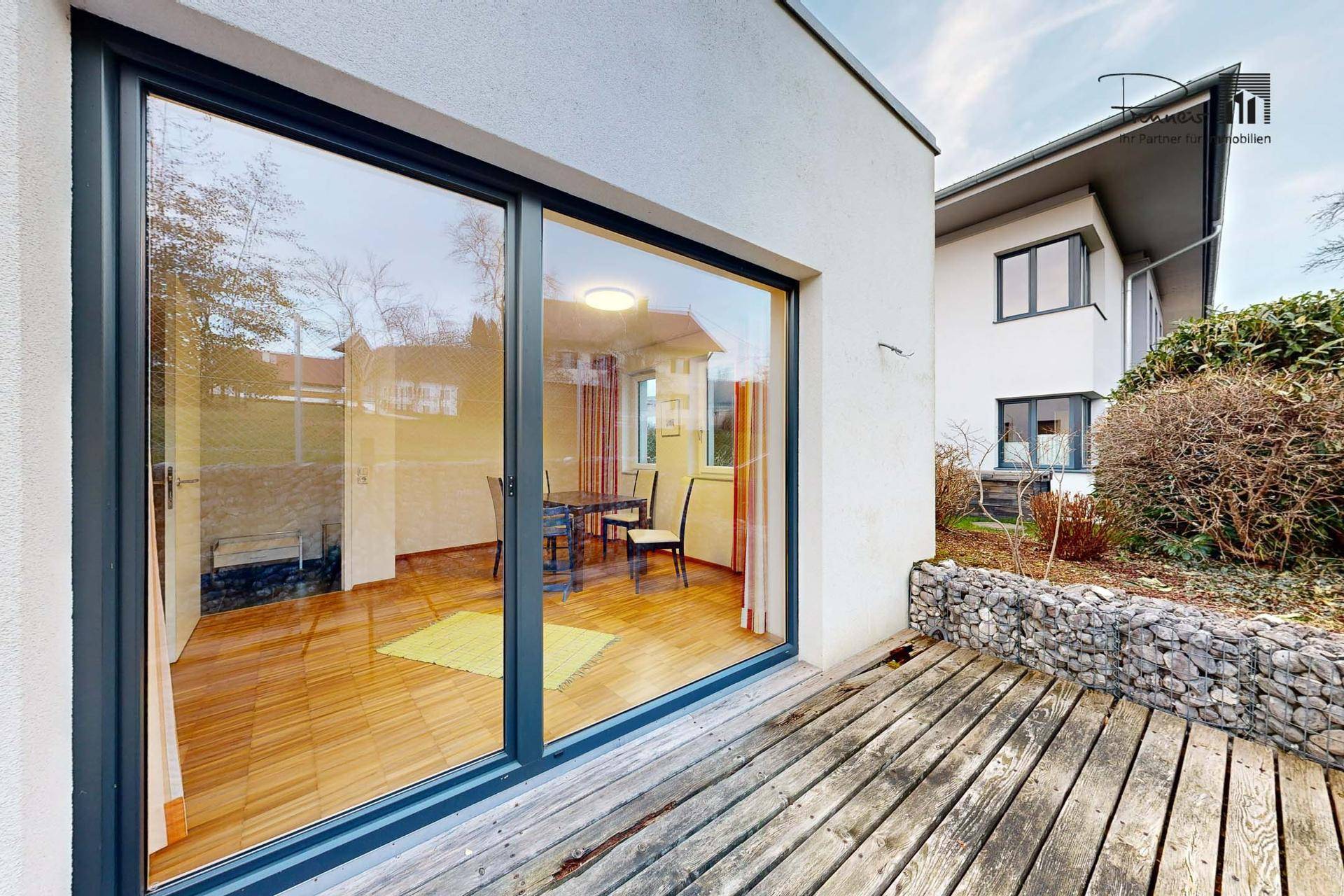 Holzterrasse mit großen Glasschiebetüren, die in einen Innenraum führen, umgeben von einer Gabionenwand.