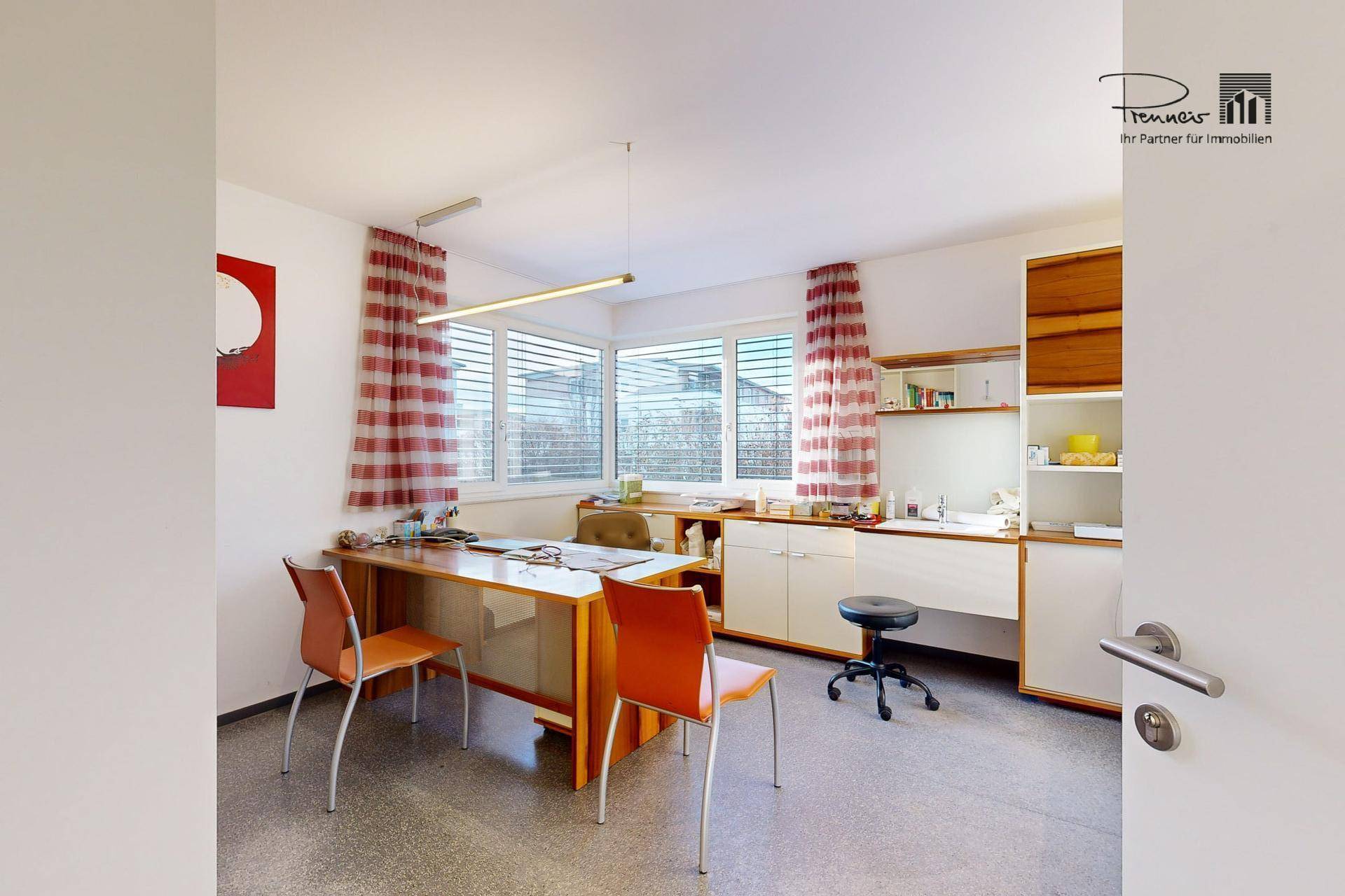 Modernes Büro oder Behandlungszimmer mit großen Fenstern, einem Schreibtisch und mehreren Stühlen in warmen Farbtönen.