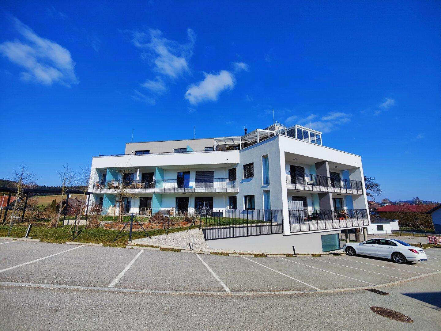 Neubau-Mehrfamilienhaus mit moderner Architektur und großem Parkplatzbereich unter strahlend blauem Himmel.