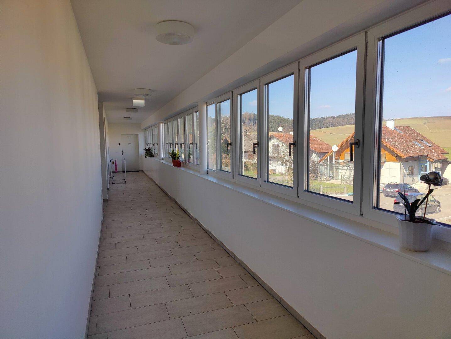 Langer, heller Flur mit vielen Fenstern und Blick auf die ländliche Umgebung und grüne Hügel.