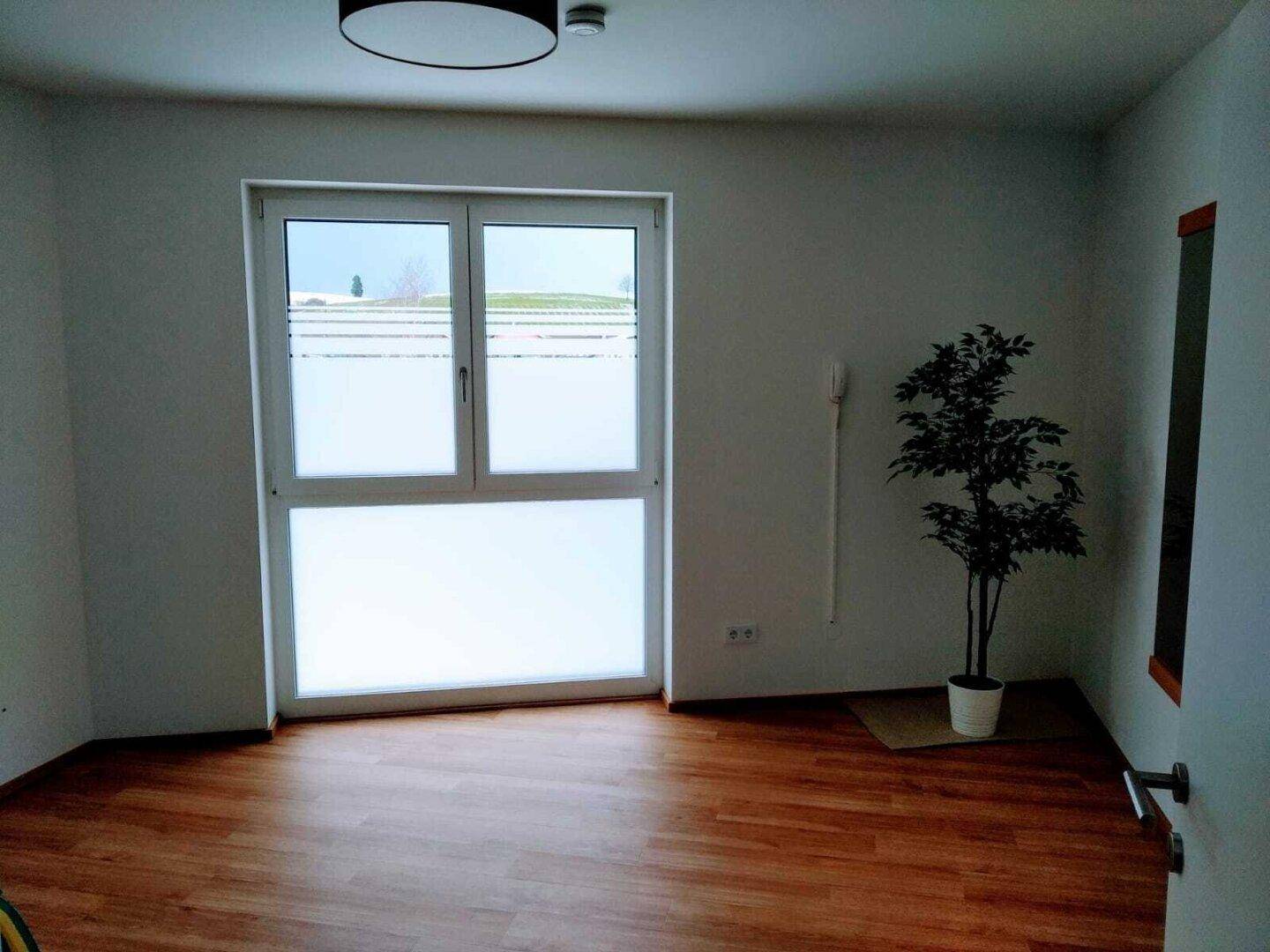 Leeres Zimmer mit Laminatboden und großem Fenster, das viel Tageslicht hereinlässt.