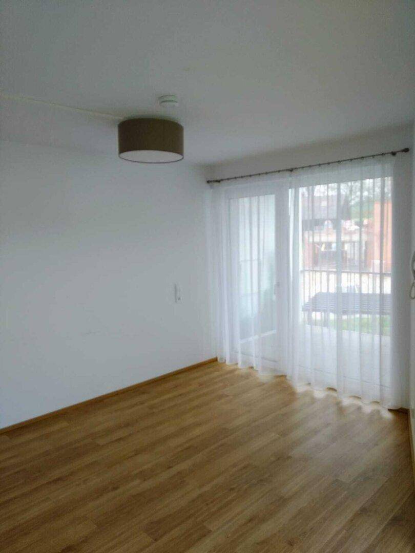 Helles Zimmer mit Laminatboden und Zugang zu einem Balkon, ausgestattet mit transparenten Vorhängen.