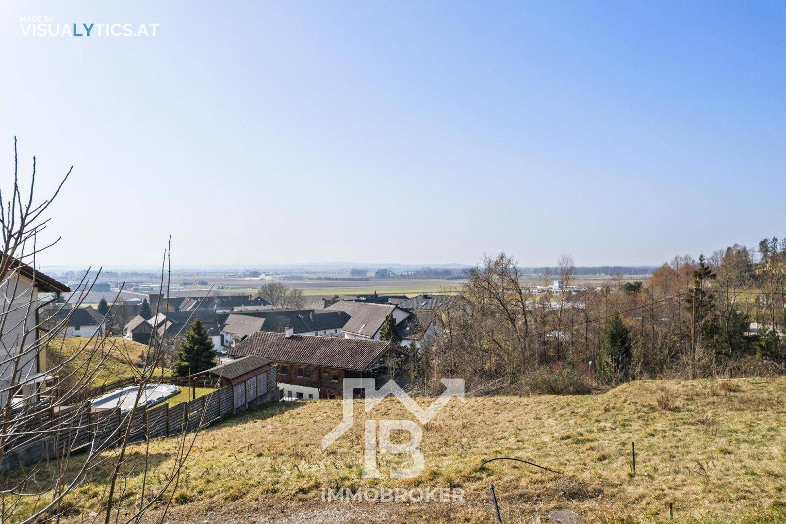 Panoramablick über eine ländliche Wohngegend mit weiten Feldern und bewaldeten Hängen am Horizont.
