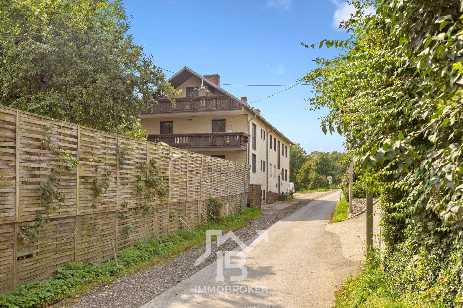 Ansicht des Mehrfamilienhauses von der Straße aus, umgeben von viel Grün und einem Holzzaun.