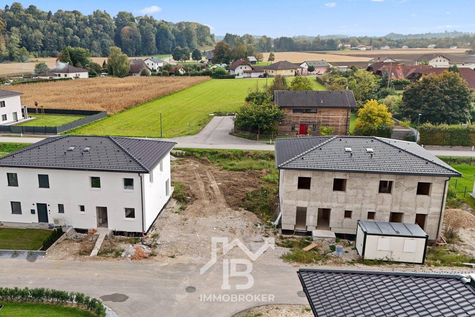 Ansicht von zwei Häusern in einer Neubausiedlung, eines fertiggestellt, das andere im Rohbau, mit ländlicher Umgebung.