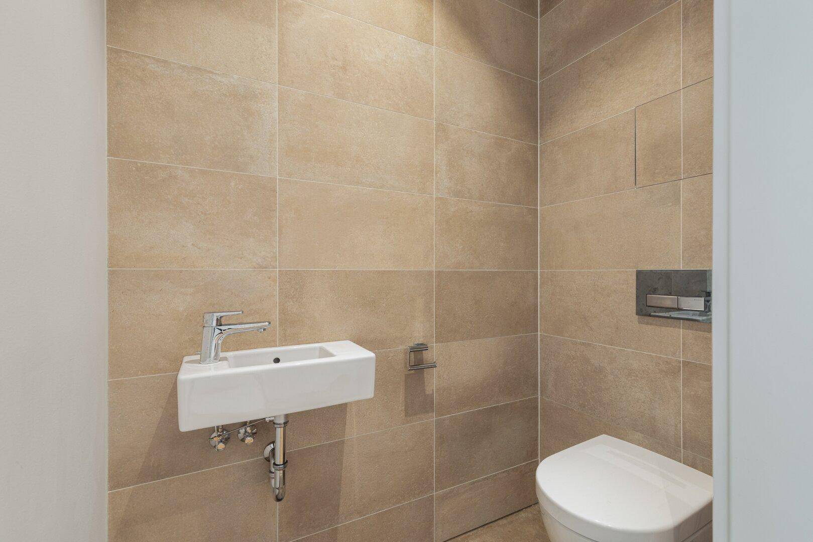 Kompakte Gästetoilette mit Wand-WC, kleinem Waschbecken und modernen beigen Fliesen.