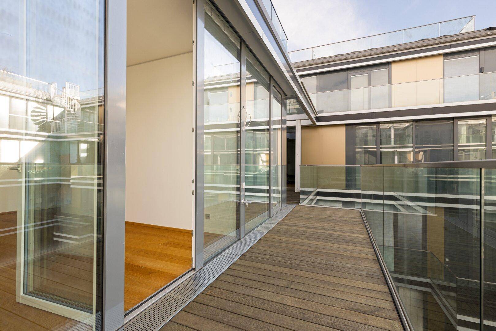 Langer Balkon mit Holzdielen und Glasbrüstung, der sich entlang der modernen Gebäudefassade erstreckt.