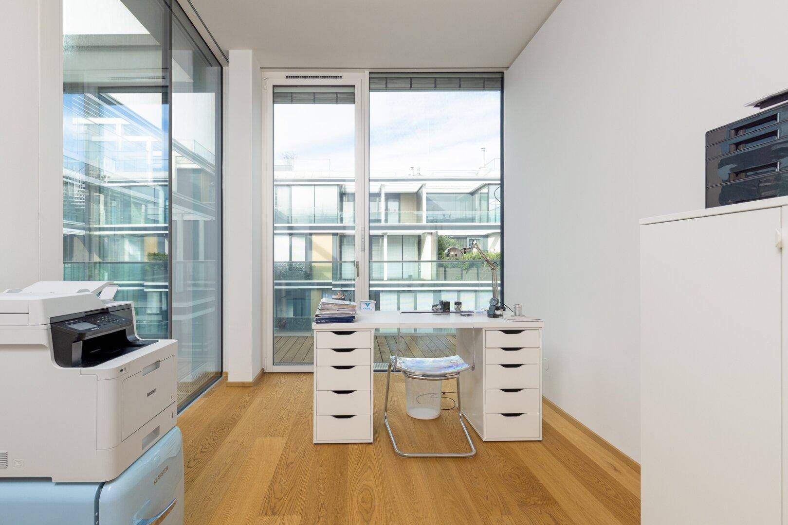 Funktionaler Büroraum mit Holzboden, großen Fenstern und Blick auf die umliegenden Balkone.