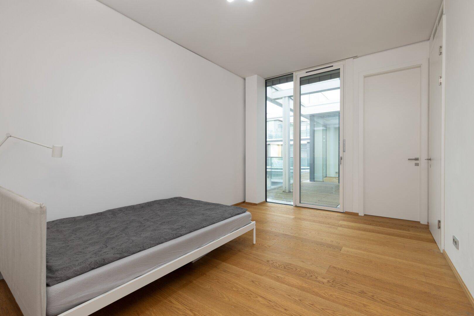 Schlichtes Schlafzimmer mit Holzboden und großen Fenstern, die Zugang zu einem Außenbereich bieten.