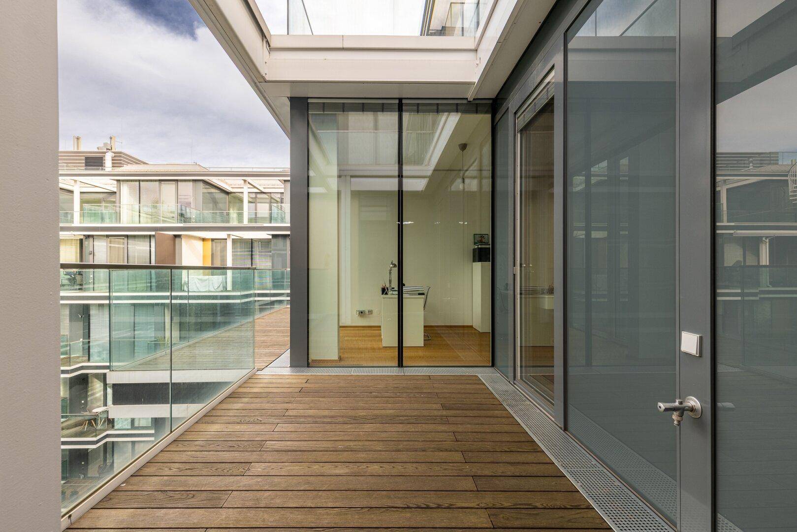 Moderner Balkon mit Holzboden und Glasgeländer, bietet einen Blick auf die umliegenden Gebäude.